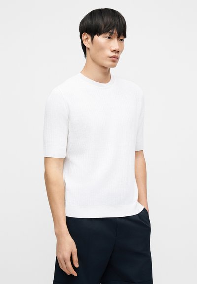 Λευκός ριμπ πλεκτός μπλουζάκι με κοντά μανίκια και στρογγυλή λαιμόκοψη, συνδυασμένο με σκούρο σορτς. Η υφή είναι λεία και ελαφριά.