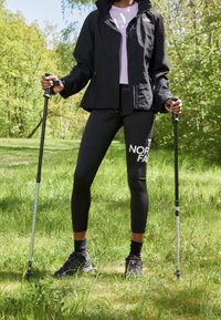 Persoon in zwarte North Face-jack, zwarte leggings met logo, met wandelstokken in de hand, staande op een graspad omringd door groene bomen.