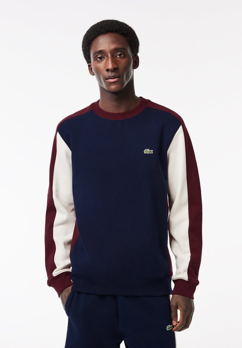 Sudadera azul marino con mangas burdeos y crema, puños acanalados, cuello redondo y pequeño logo verde en el pecho. Textura suave.