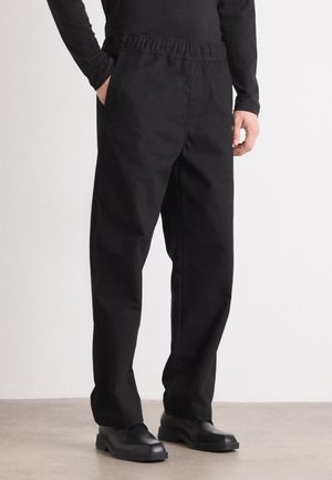 Homme portant un pantalon noir ample avec une ceinture élastique, un haut noir à manches longues, et des chaussures noires à enfiler, debout sur un sol uni.