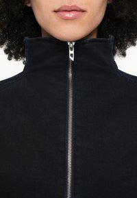 Σκούρο μπλε μπουφάν με φερμουάρ, υψηλός γιακάς, ασημένιο φερμουάρ και διακριτικές λεπτομέρειες ραφής. Λείο ύφασμα, μινιμαλιστικός σχεδιασμός.