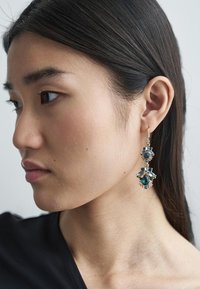 Guldfärgade hängande örhängen med blå och teal färgade ädelstenar i dropp- och blomform, med en blandning av texturer och en elegant design.