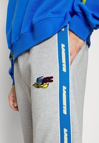 Grå sweatpants med blå sidosträck, med broderad flerfärgad Lacoste-logotyp på lårets framsida och en ficka på sidan.