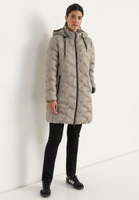 Dik grijze jas met capuchon, voorrits en twee zijzakken. Kenmerkt zich door een gewatteerd ontwerp en elastische manchetten, gecombineerd met zwarte broek en schoenen.