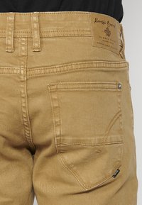 Kohvade denim-püksid tekstuuriga viimistlusega, millel on kaks tagumist taskut, tugevdusõmblused ja nahkplekk, millel on brändi logo vöökoha juures.