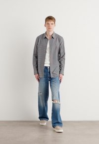 Camisa de botón gris sobre una camiseta blanca, combinada con jeans azules desgastados y zapatillas de color claro, de pie contra un fondo blanco.