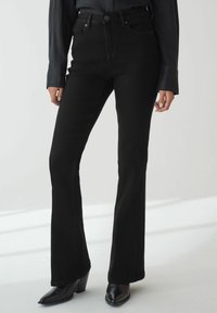 Jeans acampanados negros de cintura alta; hechos de denim, con una textura suave, costuras en contraste y bolsillos delanteros. Botas negras de tacón.