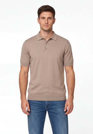 Uomo che indossa una polo beige a maniche corte e jeans blu, in piedi davanti a uno sfondo bianco uniforme.