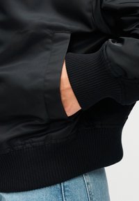 Μαύρο μπουφάν μπόνμερ με λείο ύφασμα, ριμπ μανσέτες και μέση. Περιλαμβάνει πλαϊνή τσέπη με διακριτική ραφή.