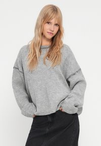 Siv sweater iz pletene tkanine z rebrasto teksturo, širokimi rokavi in črno obrobo. V kombinaciji z temno denim krilom za plasten videz.