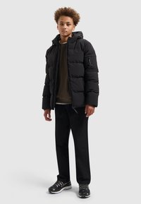 Zwarte pufferjas met capuchon, met rits aan de voorkant en zijzak, gedragen over een donkergroen sweatshirt, gecombineerd met zwarte broek en sneakers.