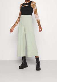 Modelka ubrana w jasnozielone szerokie spodnie, czarny bezrękawnik na brązowej koszuli z nadrukiem tie-dye z długim rękawem oraz czarne sznurowane platformy.