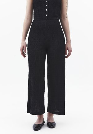 Vrouw die zwarte, hooggetailleerde, wijd uitlopende broek met textuur draagt, gecombineerd met zwarte platte schoenen, staand tegen een effen witte achtergrond.