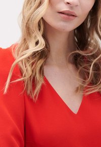 Top rojo de escote en V con una tela texturizada y mangas de corte holgado, combinado con cabello rubio ondulado hasta los hombros que enmarca la cara.