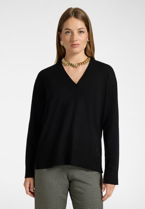 Vrouw met een zwarte V-hals trui, zwart-witte geruite broek en een stevige gouden ketting en oorbellen, staand tegen een effen witte achtergrond.