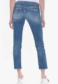 Jeans de mezclilla azul ajustados con un acabado desgastado, cinco bolsillos y un parche de cuero en la parte posterior. Los dobladillos son ligeramente recortados, dejando al descubierto zapatillas blancas.