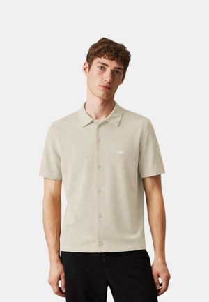 Giovane uomo con capelli corti e ricci che indossa una camicia beige a maniche corte con bottoni e pantaloni neri su uno sfondo chiaro e uniforme.