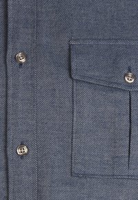 Camicia di denim con un motivo a spina di pesce blu, dotata di bottoni argentati e di una tasca frontale. Il tessuto è strutturato e resistente.