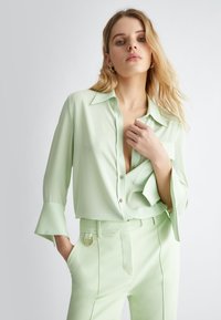 Blusa verde chiaro in tessuto morbido con chiusura a bottoni frontale, colletto a punta e maniche svasate. Abbinata a pantaloni coordinati con accessori dorati.