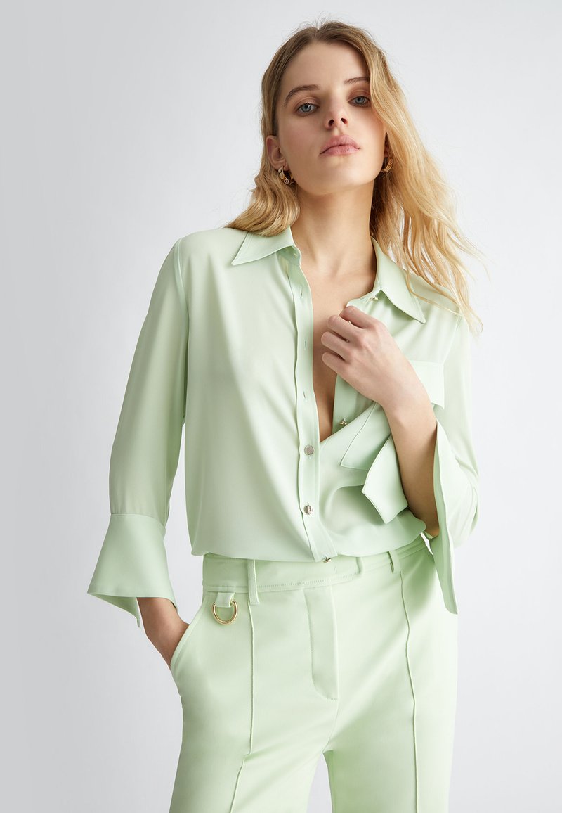 Blusa verde chiaro in tessuto morbido con chiusura a bottoni frontale, colletto a punta e maniche svasate. Abbinata a pantaloni coordinati con accessori dorati.