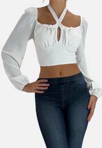White long-sleeve top with a ruffled neckline, crisscross straps, and fitted waist. Paired with dark denim high-waisted jeans.