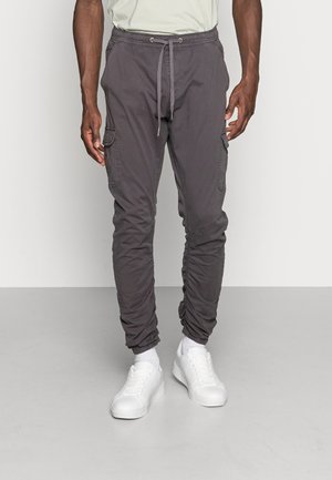 Mann trägt graue Cargo-Jogginghosen mit Kordelzug am Bund, gesammelten Bündchen, weißen Socken und weißen Turnschuhen, steht auf einem grauen Boden.