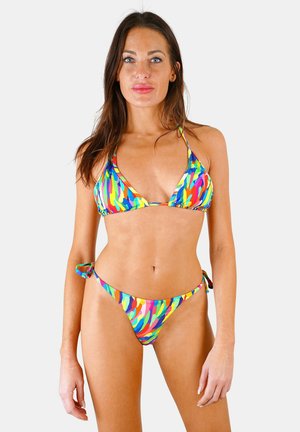 Femme aux longs cheveux bruns, debout face à l'avant, portant un bikini coloré à motif abstrait avec des liens latéraux sur fond blanc.