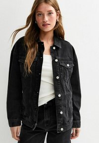 Veste en denim noir avec une coupe décontractée, dotée de boutons argentés, de poches poitrine et d'une texture légèrement délavée. Portée par-dessus un top blanc.
