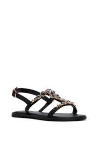 Sandal nero flat con cinturini decorati che presentano pietre trasparenti e nere, dettagli dorati e chiusura con fibbia sul cinturino alla caviglia.
