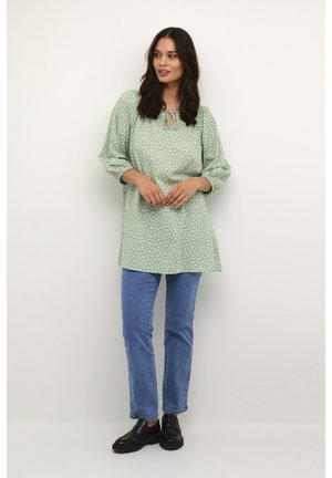 Grøn floral tunika med snøre ved halsudskæringen, lange puffede ærmer, parret med blå jeans og sorte sko. Blødt stof, afslappet pasform.