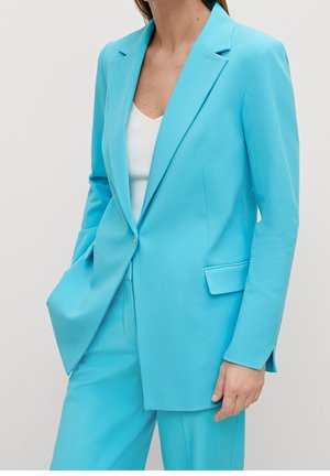 Kvinde iført en strålende blå skræddersyet blazer og matchende bukser med en hvid ribbet top, stående med den ene hånd i lommen.