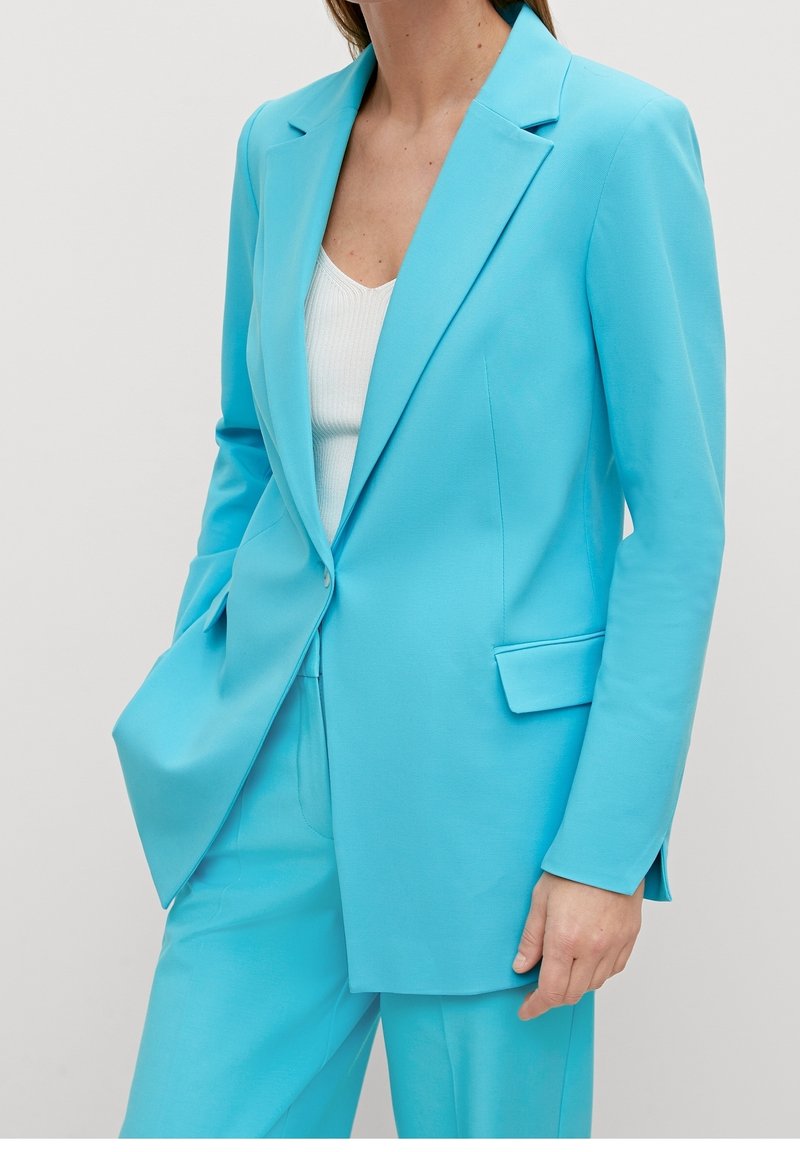 Femme portant un blazer bleu vif sur mesure et un pantalon assorti, avec un haut blanc côtelé, debout, une main dans la poche.