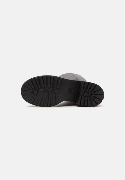 Semelle noire de botte avec des crampons profonds et un logo de marque, vue par le dessous sur un fond blanc uni.