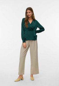 Zelena bluzica s dugim rukavima i plisiranim detaljima, uparena s bež hlačama širom nogavica i svijetložutim cipelama na remen.