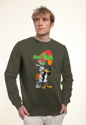 Ung mand iført olivengrøn sweatshirt med Bugs Bunny og Daffy Duck, der spiller basketball, samt "Space Jam"-logo.