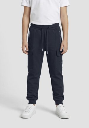 Person wearing black cargo jogger pants with zip and flap pockets, white sneakers, and a white short-sleeve shirt, standing against a plain background.