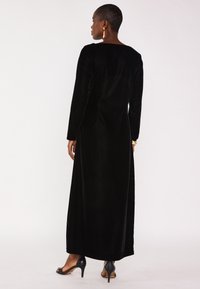 Robe longue en velours noir à manches longues, avec une silhouette droite et un dos décolleté. Le mannequin porte des talons noirs et des accents de bijoux en or.