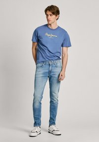 Camiseta de color azul claro con logo, combinada con jeans azules desgastados. El modelo lleva zapatillas blancas y negras. Diseño simple, casual y ajustado.