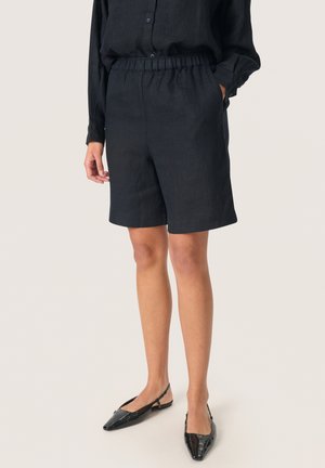 Personne portant un short noir taille élastique et un haut noir à manches longues, associé à des ballerines slingback noires à bout pointu sur un fond clair.