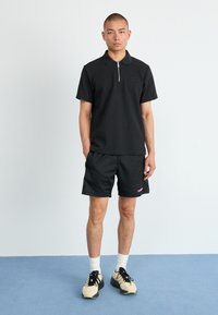 Polo noir à manches courtes avec une fermeture éclair sur le devant, accompagné de shorts noirs avec une marque rose, porté avec des chaussettes blanches et des baskets.