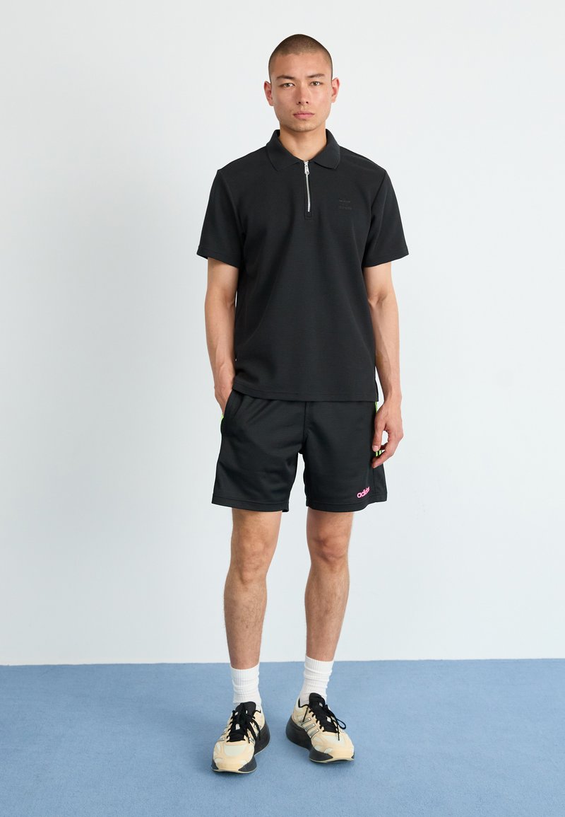 Polo noir à manches courtes avec une fermeture éclair sur le devant, accompagné de shorts noirs avec une marque rose, porté avec des chaussettes blanches et des baskets.