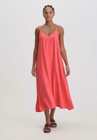 Koralli maksimekko, jossa on ohuet olkaimet, V-pääntiä, sileä tekstuuri, kevyt kangas ja virtaava siluetti. Yhdistetty ruskeisiin sandaaleihin.