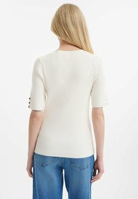 Bílý žebrovaný pletený top s krátkými rukávy a černými knoflíkovými detaily na manžetách, kombinovaný s modrými džíny, minimalistický design.