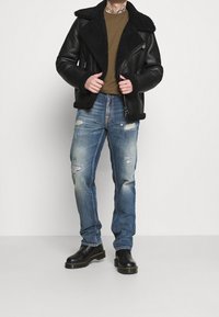 Veste en cuir noir doublée de peau avec un grand col, chemise verte en dessous, jean bleu usé, et bottines noires.