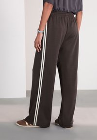 Braune, weit geschnittene Hose mit weißen seitlichen Streifen, elastischem Bund und glattem Stoff. Kombiniert mit braunen und weißen Schuhen.