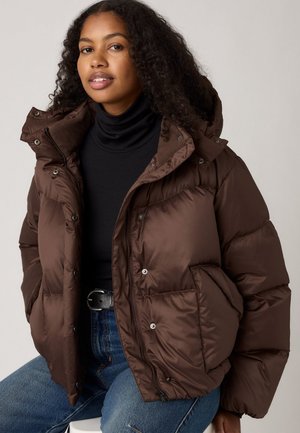 Bruine pufferjack met een capuchon, met een glanzende afwerking, drukknoopsluiting en zijzakken, gedragen over een zwarte turtleneck en blauwe jeans.