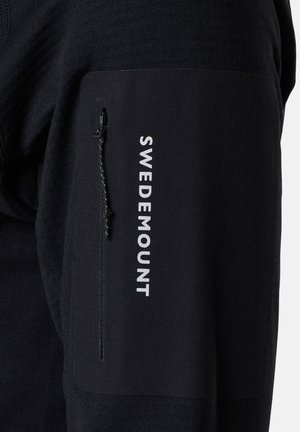 Musta neule, jossa on vetoketjullinen tasku hihassa, esittelee "SWEDEMOUNT" -tekstiä valkoisilla kirjainmerkeillä. Pehmeä kangas ja istuva muotoilu.