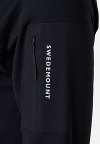 Sort langærmet pullover med tekstureret stof. Har en lynlåslomme på venstre arm og logoet "SWEDEMOUNT" i hvidt på lommen.