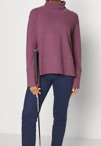 Langärmliger Pullover mit hohem Kragen aus tiefpurpur farbenem Stoff und lockerer Passform. Kombiniert mit dunkelblauen Hosen und hält einen Golfschläger.