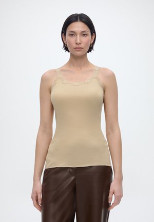 Γυναίκα με κοντά σκούρα μαλλιά, φορώντας αμάνικο μπεζ ριμπ τοπ με δαντελένια λεπτομέρεια και σκούρο καφέ δερμάτινο παντελόνι, στέκεται μπροστά σε απλό λευκό φόντο.